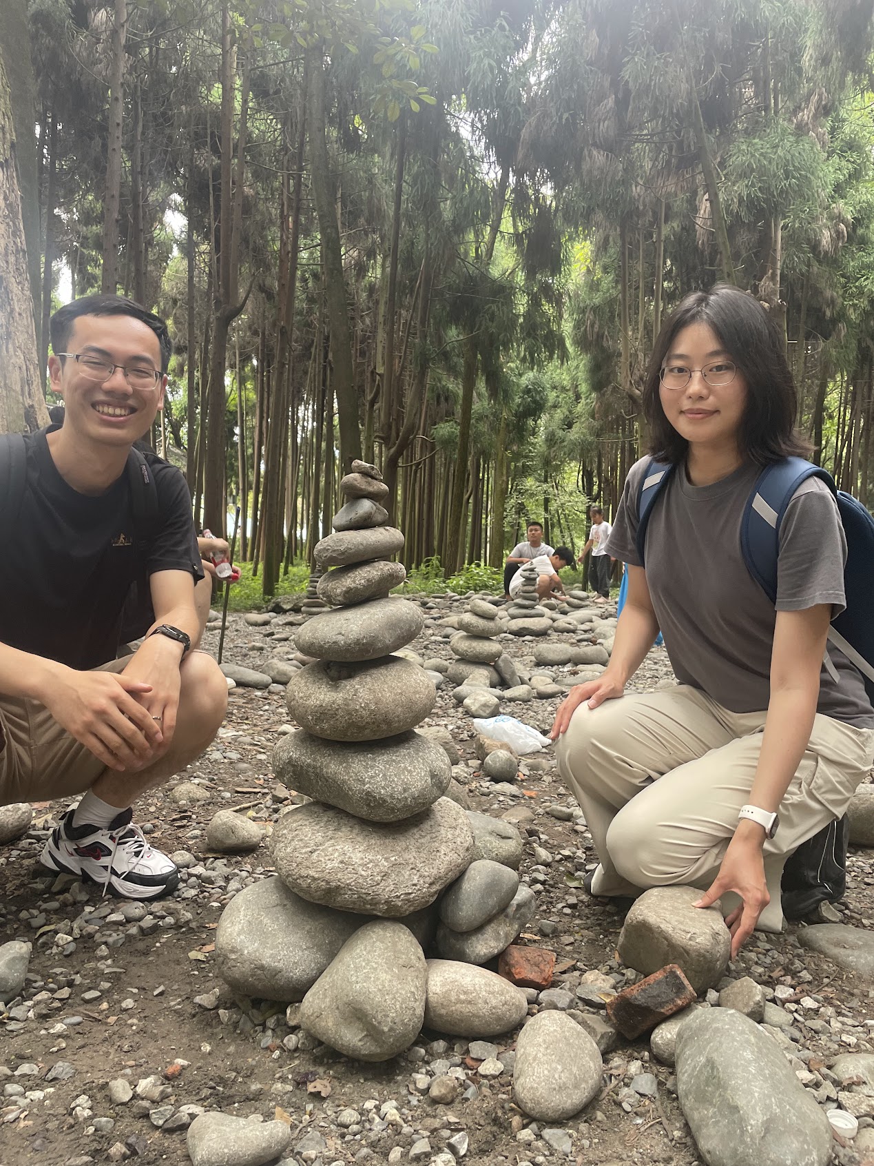 Two CANN lab members crouching beside a balanced stone sculpture in a wooded area.